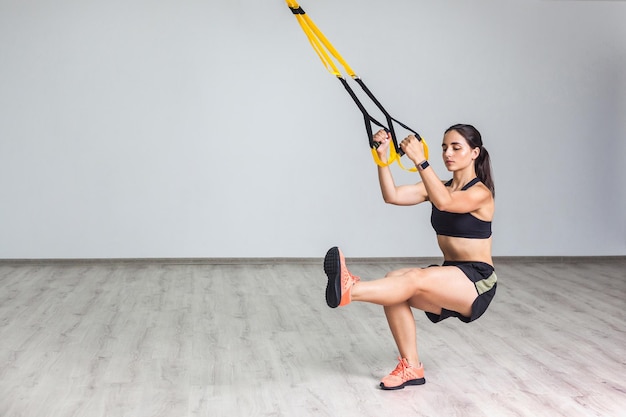 Portrait of young athletic woman in sportswear doing squat exercise, training legs and glutes muscular with fitness straps in the gym. Functional training. Isolated, trx concept, white wall background