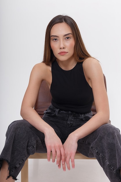 Portrait of young attractive asian woman with long brown hair in black clothes, isolated on white  wall. skinny pretty female sitting on chair on cyclorama. model tests of beautiful lady