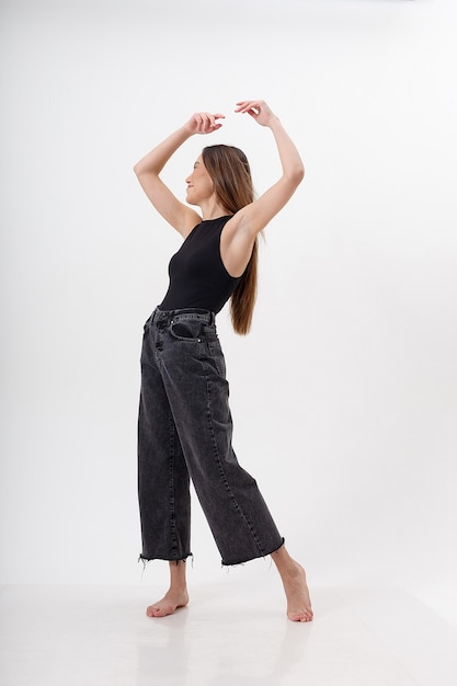 Ritratto di giovane donna asiatica attraente con capelli lunghi in tuta nera, jeans, isolato sul muro bianco. femmina graziosa magra in posa sul ciclorama. prove modello di bella signora