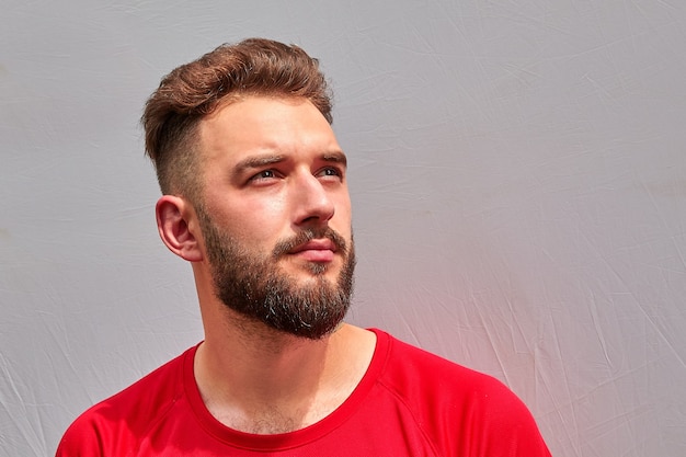 Portrait of young attractive bearded caucasian man with serious look in red T-shirt on grey background.