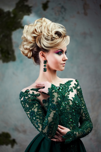 Portrait of young attractive blonde woman in a beautiful green dress. Textured background, interior. Luxury hairstyle