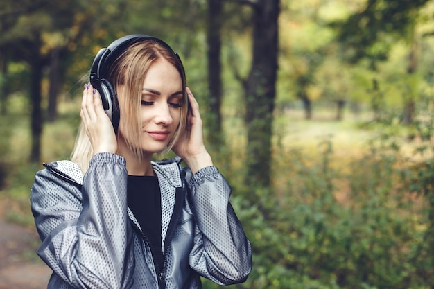 Ritratto di giovane donna bionda attraente su un parco cittadino, ascoltando musica in cuffia