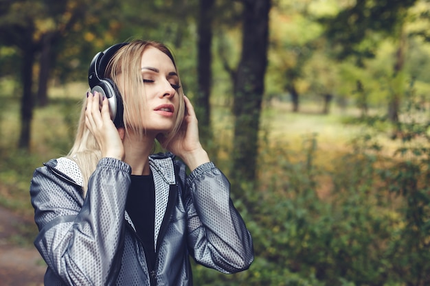 ヘッドフォンで音楽を聞いて、都市公園の若い魅力的なブロンドの女性の肖像画