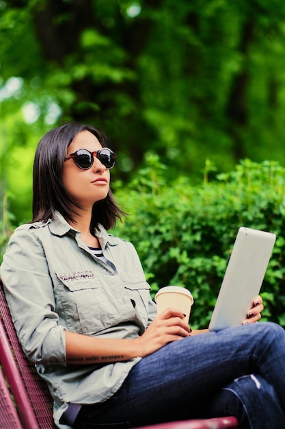 Portrait of young attractive brunette female in sunglasses holds tablet PC drinks coffee in a green summer park.