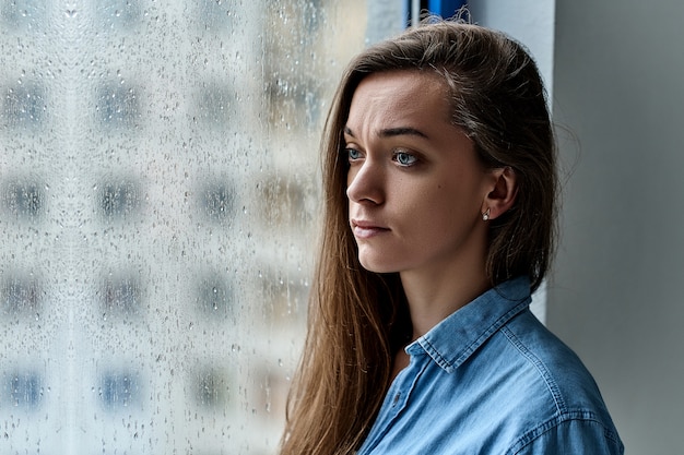長い髪と雨の秋の天候で雨滴と窓の近くに一人で立っている悲しい目を持つ若い魅力的なブルネット孤独な憂鬱物思いにふける白人女性の肖像画