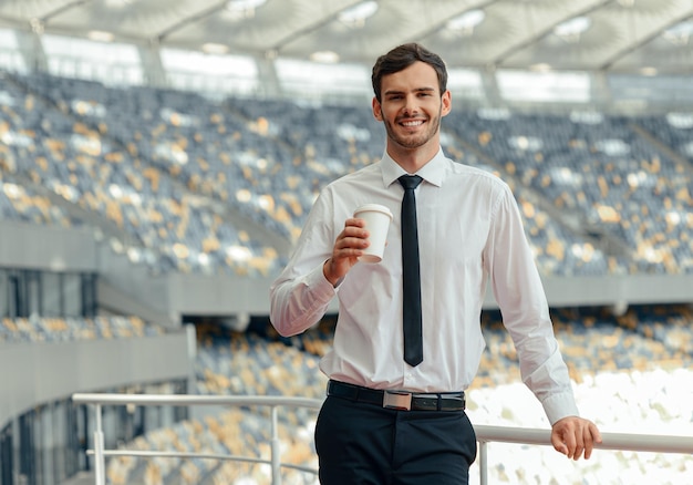 Portrait of young attractive businessman dressed in shirt standing on observation deck of stadium with cup of coffee and looking at camera