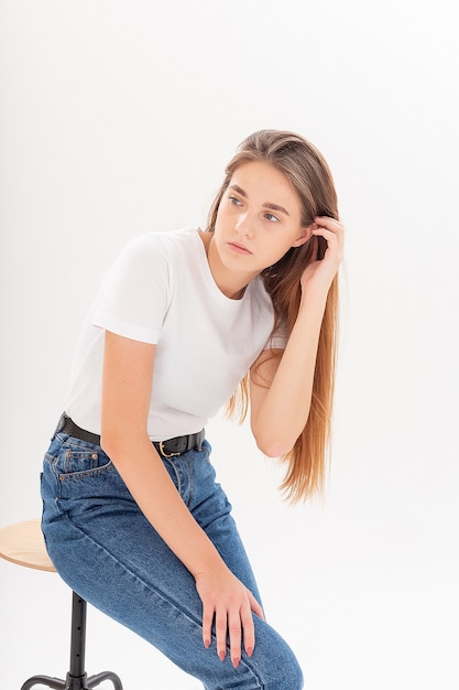 Portrait of young attractive caucasian woman with long hair in t-shirt and blue jeans isolated on white background. skinny pretty female sitting on stool at studio. model tests of beautiful lady