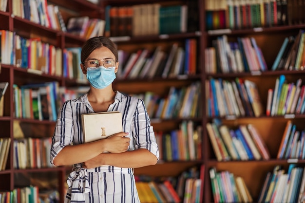 Portrait of young attractive college girl standing in library with face mask on holding a book. Studying during covid 19 concept.