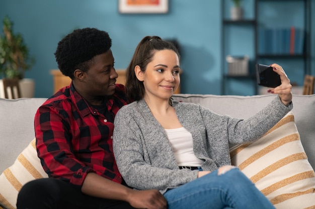 Portrait of the young attractive couple posing funny while making selfies on the smartphone in the cozy living room Indoors