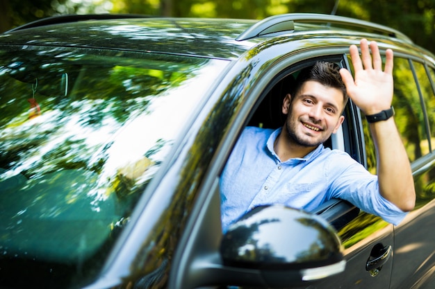 Portrait of young attractive handsome brunette man driving car and greeting somebody with hand