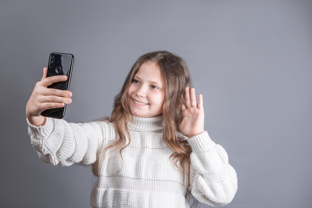 Portrait of a young attractive little girl with blond long flowing hair in a white sweater hold mobile phone takes a selfie talks on video communication 