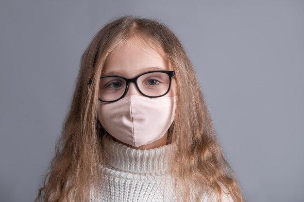 Portrait of a young attractive little girl with blond long flowing hair in a white sweater in medical protective mask on a gray studio background. Place for text. Copy space.