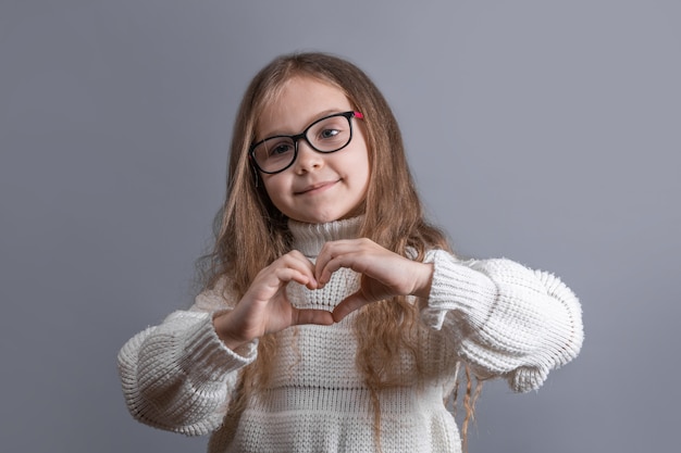 Portrait of a young attractive little girl with blond long flowing hair in a white sweater smiling shows heart hands on a gray studio background. Place for text. Copy space.