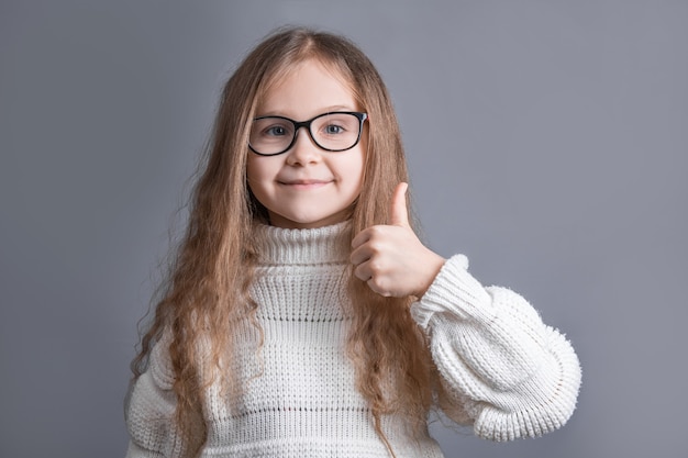 白いセーターの笑顔で金髪の長く流れる髪を持つ若い魅力的な少女の肖像画は、灰色のスタジオの背景で歌うように、親指を上に表示します。テキストの場所。スペースをコピーします。