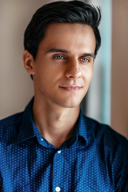 Foto ritratto di giovane uomo attraente con gli occhi azzurri e i capelli scuri, con indosso una camicia blu che guarda lontano, sorridente mentre posa