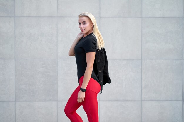 Portrait of young attractive sporty girl or model wearing black sportswear, trendy red leggings. Outdoor shot on grey wall background.