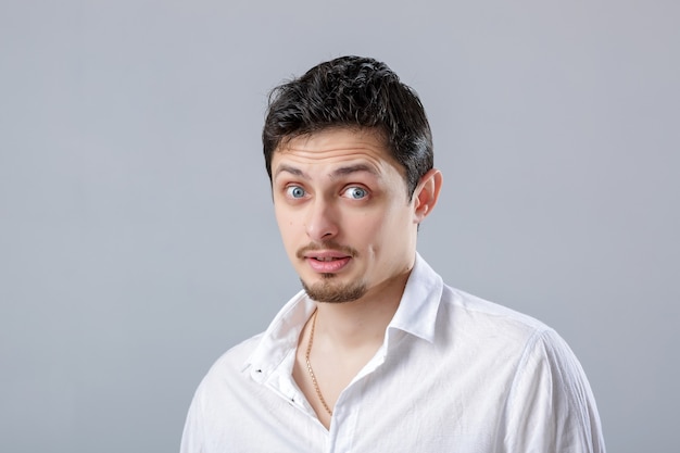 Portrait of young attractive surprised brunette male in white shirt on gray background.