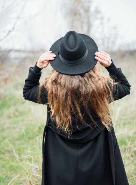 Photo portrait of young attractive woman in black coat and hat autumn landscape dry grass back view