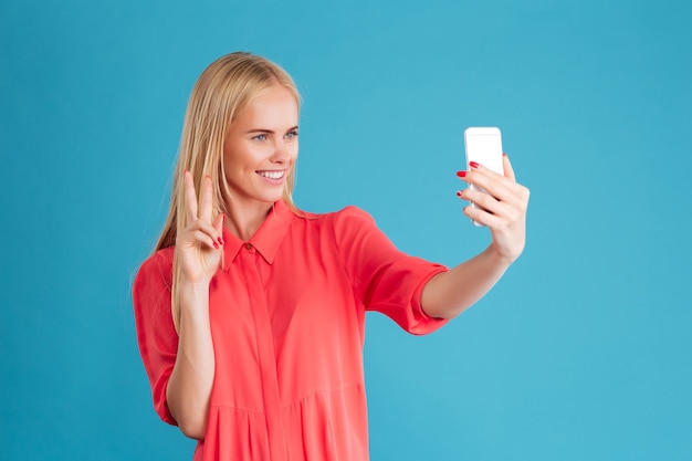 Portrait of a young attractive woman making selfie photo on smartphone over blue wall