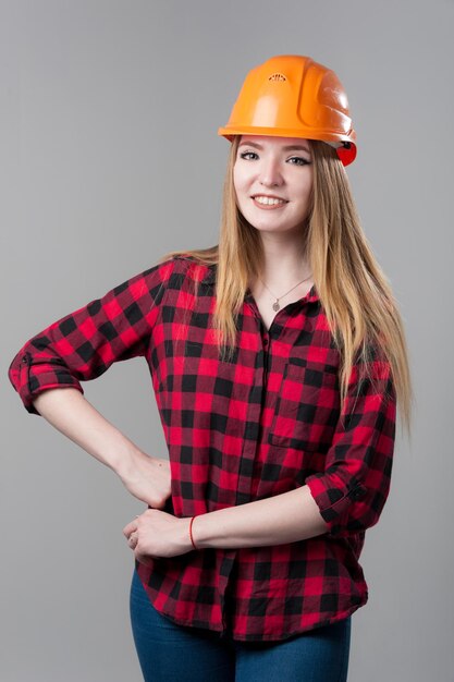 Портрет молодой привлекательной женщины со светлыми волосами в оранжевом шлеме и клетчатой рубашке на нейтральном сером фоне.