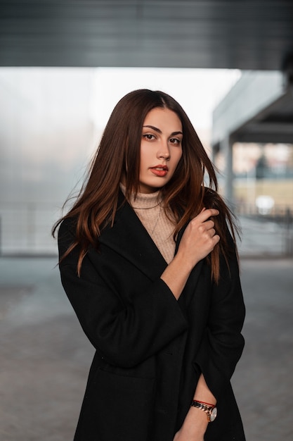 Portrait young attractive woman with luxurious long brown hair with sexy lips with natural makeup in an elegant black coat near a luminous modern building. Cute girl fashion model on the street.