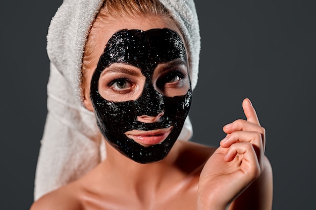 Portrait of a young attractive woman with a towel on her head\
with a cleansing black mask on her face on a gray wall.