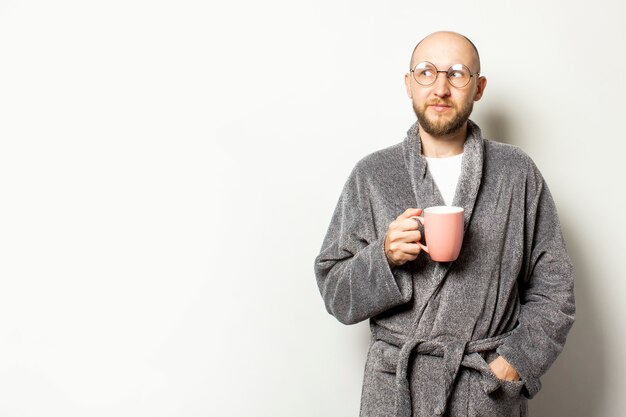 Portrait of a young bald man with a beard in a dressing gown and glasses holding a cup of coffee