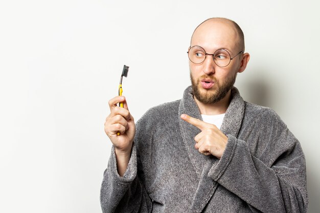 Portrait of young bald man with a beard in a dressing gown and glasses with a surprised face and points a finger at a toothbrush