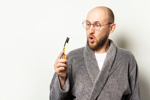 Portrait of a young bald man with a beard in a dressing gown and glasses with a surprised face and a toothbrush in his hand