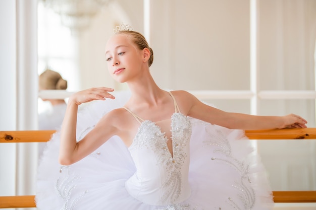 Portrait of a young ballerina in a beautiful white tutu near a ballet barre in a large beautiful white hall.