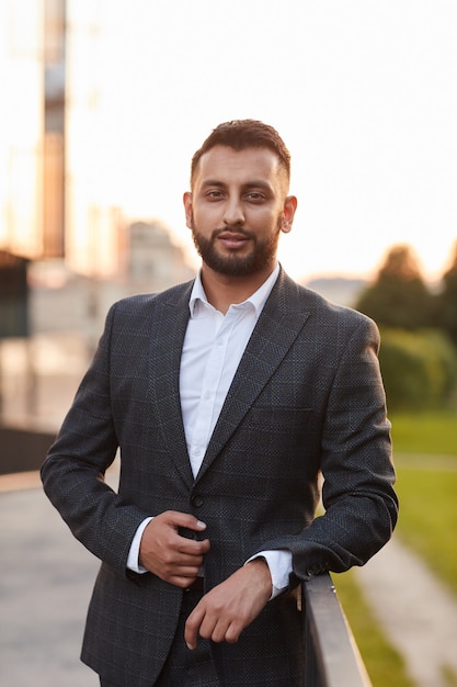 Portrait of young bearded businessman in black suit looking at camera standing outdoors