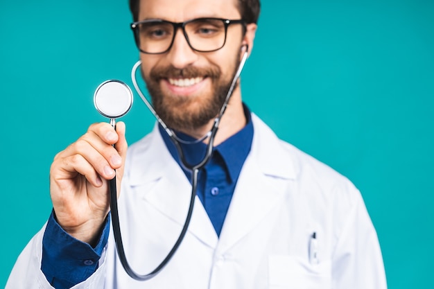 Портрет молодого бородатого доктора человека со стетоскопом на шее в медицинском пальто, стоящем на изолированном синем фоне.