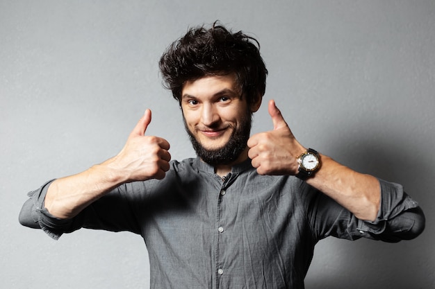 Portrait of young bearded guy with disheveled hair, show thumbs up on of grey.