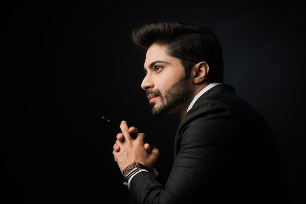 Portrait of young bearded Indian businessman against black background, moody lighting