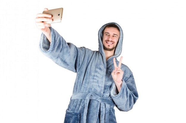 Portrait of young bearded man in blue bathrobe shows victory gesture and makes selfie