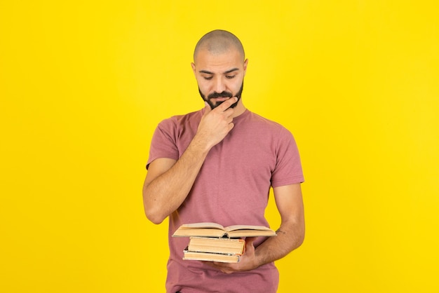 Portrait of a young bearded man holding books over yellow wall . 