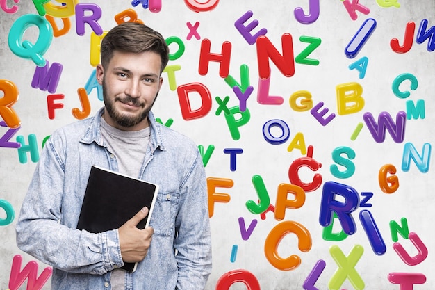 Photo portrait of a young bearded man in a jeans shirt holding a large black copybook and standing near a concrete wall with colorful letters on it mock up