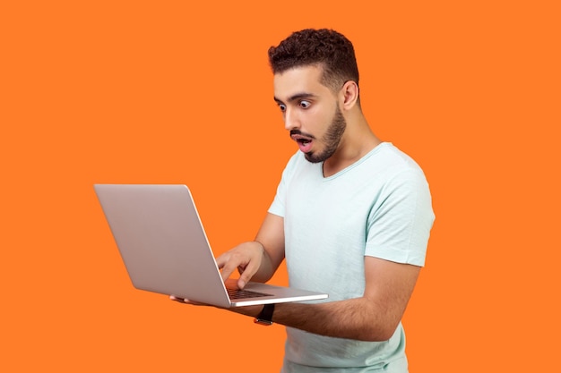 Portrait of young bearded man shocked by message on laptop having troubles while working on computer program scared startled face with open mouth and big eyes indoor isolated on orange background