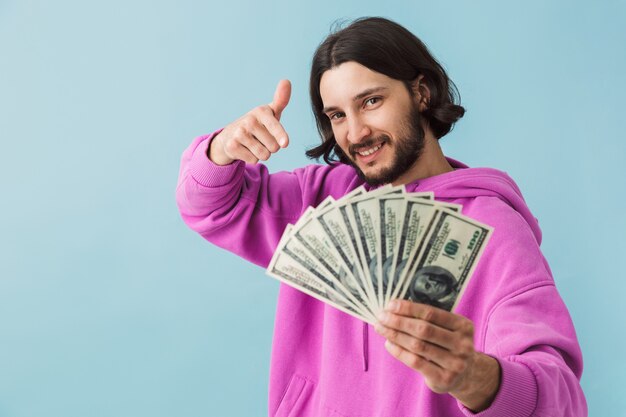 Photo portrait of a young bearded man wearing casual clothes standing isolated over wall, showing money banknotes, pointing finger