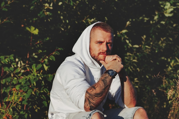 Portrait of young bearded man with tattoos in white jacket in countryside or in park. Male in casual clothes spends evening in nature. Concept of style, walking in fresh air and unity with nature