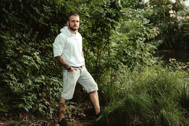 Portrait of young bearded man with tattoos in white jacket in countryside or in park. Male in casual clothes spends evening in nature. Concept of style, walking in fresh air and unity with nature