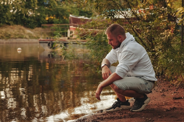 Portrait of young bearded man with tattoos in white jacket in countryside or in park. Male in casual clothes spends evening in nature. Concept of style, walking in fresh air and unity with nature