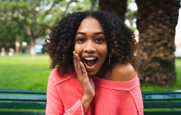 Portrait of young beautiful afro american woman with surprise expression sitting on bench in the park. Outdoors.