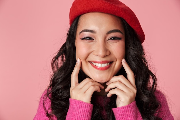 Portrait of young beautiful asian girl wearing beret showing her smile with fingers isolated on pink