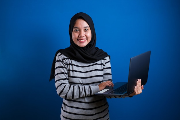 Portrait of young beautiful Asian muslim woman smiling while holding laptop, sucessfull student against blue background