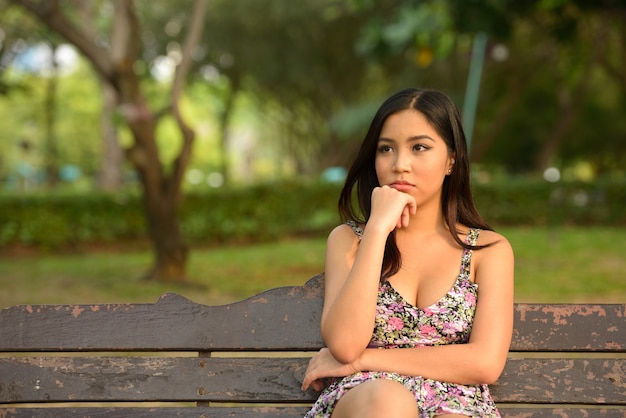 Portrait of young beautiful Asian woman sitting and thinking at the park