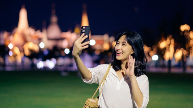 バンコク タイの夜景ポイントでエメラルド仏寺院やワット プラケオで旅行中に笑顔とスマート フォンを使用して若い美しいアジア女性の肖像画