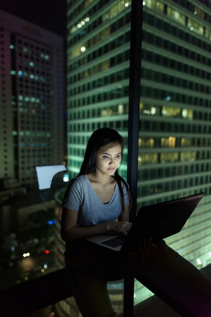 Portrait of young beautiful Asian woman using laptop against glass window with view of the city at night