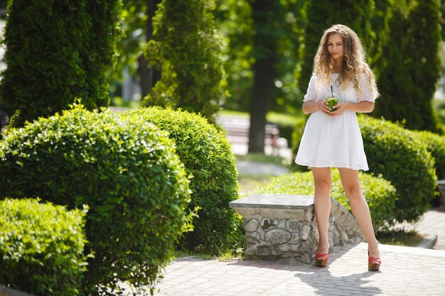 Portrait of a young beautiful attractive woman outdoors in the summer with a glass of ice cold juice or drink. Pretty girl outside with fresh mojito full height