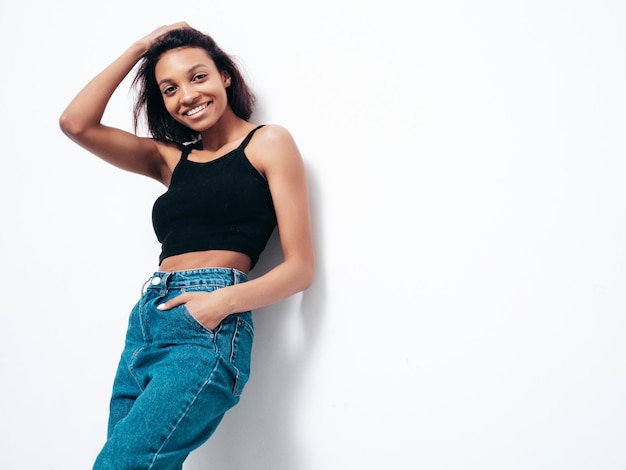 Portrait of young beautiful black woman Smiling model dressed in summer jeans and black top clothes Sexy carefree female posing near white wall in studio Tanned and cheerful